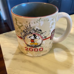 May include: A ceramic coffee mug with a white handle and a beige exterior featuring a Disneyland 2000 design. The design includes Mickey Mouse, a map, and various park attractions. The interior is green.