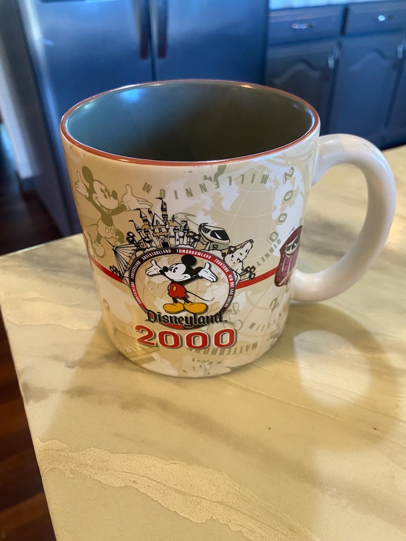 May include: A ceramic coffee mug with a white handle and a beige exterior featuring a Disneyland 2000 design. The design includes Mickey Mouse, a map, and various park attractions. The interior is green.