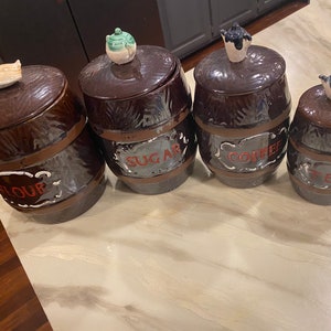 May include: Set of four brown ceramic canisters with lids shaped like teapots. The canisters are decorated with a brown and white woodgrain pattern and have labels that read "Flour", "Sugar", "Coffee", and "Tea".