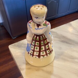 May include: A vintage ceramic salt shaker in the shape of a woman. The shaker is painted with a white dress, a brown and green checkered apron, and a red head covering. The base is yellow.
