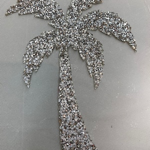 Puede incluir: Un diseño de palmera plateado hecho de pedrería. La palmera es un motivo popular para la decoración del hogar y la moda.