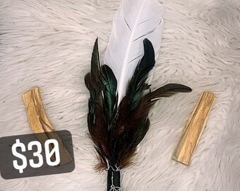 May include: A white feather smudge stick with black and green feathers, a black handle with a silver pentagram charm, and a clear crystal. The smudge stick is on a white fluffy surface with a brown stick of sage and a price tag that says £30.