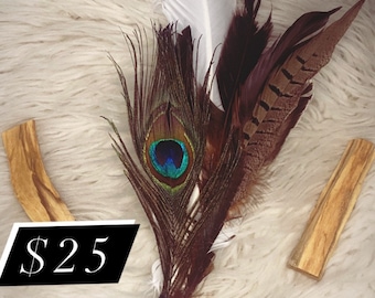 May include: A smudge stick featuring a mix of peacock, white and brown feathers. The stick is wrapped in black fabric and has a clear crystal at the end. The smudge stick is resting on a white fluffy surface. £25