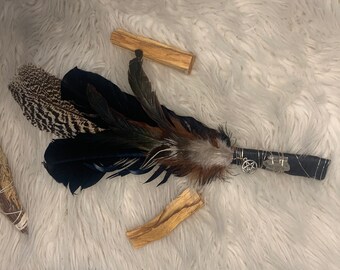 May include: A black and brown feather smudge stick with a silver pentacle charm. The smudge stick is wrapped in black fabric and has a wooden handle. There are two pieces of palo santo wood in the background.