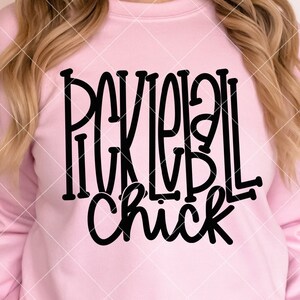 以下が含まれることがあります： ピンクのトレーナーに、黒い文字で「Pickleball Chick」と書かれています。
