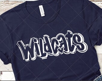 May include: A navy blue t-shirt with the word "Wildcats" in white outlined letters. The text is in a graffiti style font. The shirt is laying on a wooden surface with a pair of jeans and a flip-flop.