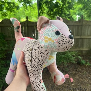 May include: A handmade stuffed animal dog with a patchwork design. The dog features a pink body with floral and dotted fabric accents. It has black button eyes and a button closure on its side. The dog is held in front of a blurred green background.