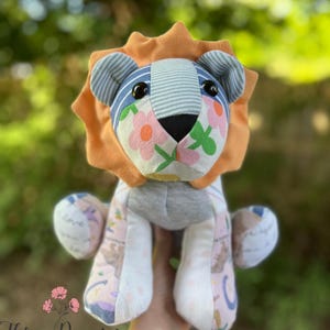 May include: A handmade lion plush toy with a patchwork design. The lion features an orange mane, blue and white striped ears, and a face adorned with floral fabric. The body is a mix of patterned fabrics, and the toy is held in a hand.