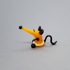 May include: A small glass figurine of a mouse. The mouse is yellow with black ears, a black nose, and a black tail. The mouse is sitting on its hind legs with its front paws raised.