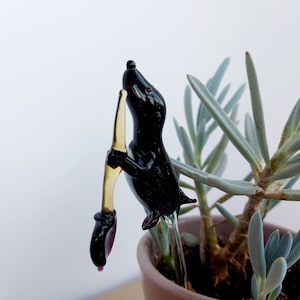 May include: A black glass animal figurine of a mouse climbing a yellow glass stick. The figurine is in a pot with a green succulent plant.