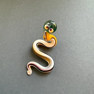Op de afbeelding: Een handgemaakte glazen slangenfiguur met een groene kop, gele nek en een zilver-roze lichaam. De slang heeft paarse ogen. Het beeldje is een decoratief item.