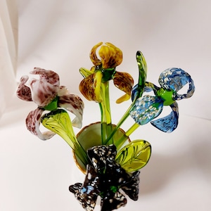 Può includere: Un mazzo di quattro fiori di iris di vetro in un vaso bianco. I fiori sono in tonalità di marrone, giallo, blu e nero con steli verdi.