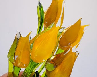 May include: A bouquet of five handcrafted glass tulips featuring vibrant yellow petals and translucent green stems and leaves. The glass flowers are arranged in a cluster, showcasing smooth, reflective surfaces and clear, defined shapes.