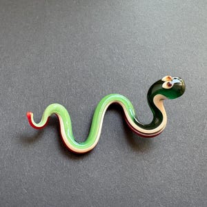 May include: A glass snake figurine with a green body, white and red stripes, and a dark green head. The snake is curled in a wavy pattern, with a red tip on its tail. The figurine is on a grey surface.