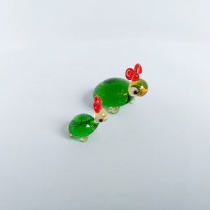 May include: Two green glass turtle figurines with red bows on their heads. The larger turtle has yellow eyes and the smaller turtle has white eyes.