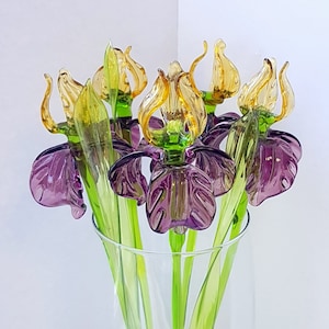 May include: A bouquet of six glass irises in a clear glass vase. The irises have purple petals, green stems, and yellow-gold accents.