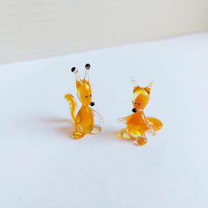 May include: Two small glass figurines of squirrels. The squirrels are orange and have black eyes. They are standing on a white surface.