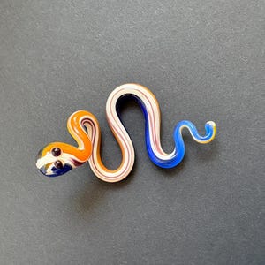 Peut inclure: Figurine de serpent en verre coloré. Le serpent est principalement orange, blanc et bleu, avec des accents bruns pour les yeux. Le serpent mesure environ 15 cm de long et a une forme incurvée et sinueuse.