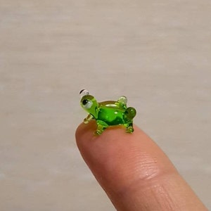 以下が含まれることがあります： 白い目をした小さな緑色のガラス製のカエルの置物。カエルは指の上に座っています。