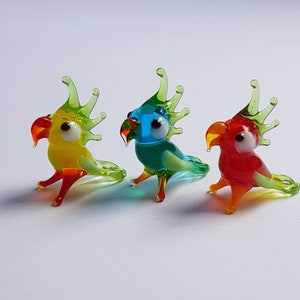 May include: Three glass parrot figurines, each with a different colour scheme. The first parrot is yellow with green accents, the second is blue with green accents, and the third is red with green accents.