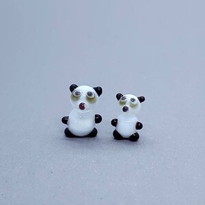 Puede incluir: Dos figuritas de panda de vidrio blanco con ojos y orejas negros. El panda más grande está de pie con la boca abierta, mientras que el panda más pequeño está de pie con la boca cerrada.