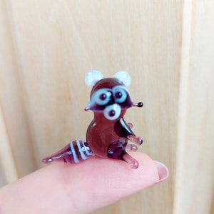 May include: A small glass figurine of a raccoon with purple and white stripes. The raccoon has white ears and black eyes.