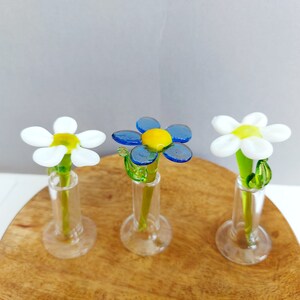 Può includere: Tre vasi di fiori di vetro con fiori di vetro bianchi e blu. I fiori sono in un piccolo vaso di vetro con una base rotonda. I vasi sono su una superficie di legno.
