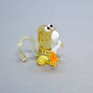 Könnte beinhalten: Eine kleine Glasfigur eines Affen, der mit gekreuzten Beinen sitzt und eine Banane in den Händen hält. Der Affe ist gelb mit weißen Augen und einem weißen Mund. Die Banane ist orange und gelb.