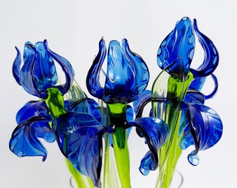 May include: A bouquet of blue glass irises with green stems in a clear glass vase. The irises are arranged in a fan shape, with the tips of the petals pointing upwards.