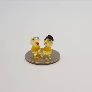 May include: Two small glass figurines of yellow birds with black eyes. One bird is wearing a black hat. The figurines are sitting on a silver US quarter.