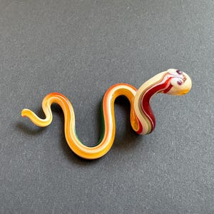 May include: A glass snake figurine with a curved body. The snake has a cream-coloured head with red and white stripes, and a body with orange, yellow, and green stripes. The figurine is on a grey surface.