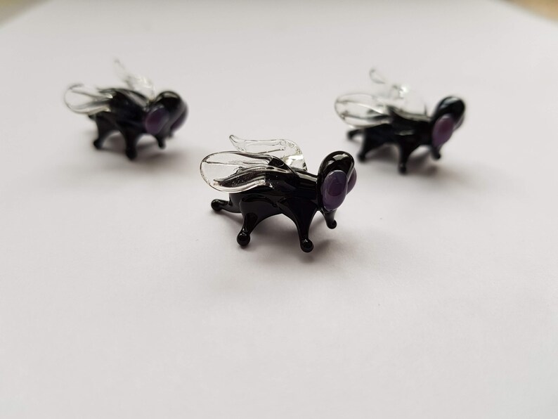 May include: Three black glass flies with clear wings and purple eyes. The flies are sitting on a white surface.