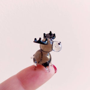 May include: A small glass moose figurine with black antlers and brown glass body. The moose is standing on a finger.
