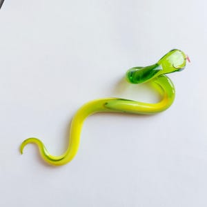 以下が含まれることがあります： 手作りのガラス製のヘビの置物。ヘビの体は黄色と緑のグラデーションで、鮮やかな緑色の頭と小さなピンクの舌があります。ヘビは曲がった形に巻かれています。