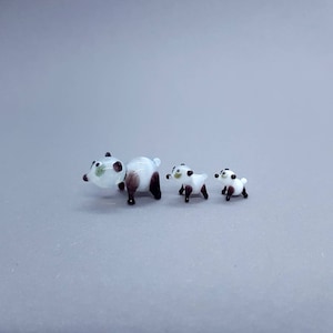 Puede incluir: Tres figuritas de panda de vidrio, cada una con ojos negros y un cuerpo blanco con marcas negras. El panda más grande está de pie sobre sus cuatro patas, mientras que los dos pandas más pequeños están sentados.