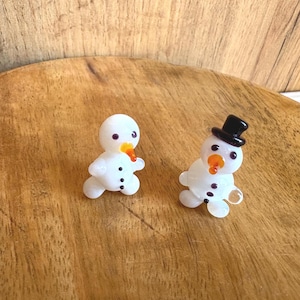 May include: Two small, white snowman figurines with black button details, orange carrot noses, and purple eyes. One snowman wears a black top hat. The snowmen are on a wooden surface.