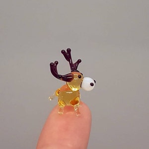 Op de afbeelding: Een miniatuur glazen rendierfiguur met een amberkleurig lichaam, donkerbruin gewei en een wit en zwart oog. De miniatuursculptuur staat op een vingertop tegen een neutrale achtergrond.