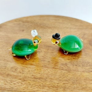 May include: Two small glass turtle figurines, one wearing a white bow and the other a black top hat, on a wooden surface.