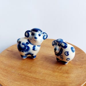Puede incluir: Dos pequeñas figuritas de ovejas de cerámica pintadas con un patrón azul y blanco. Las ovejas están de pie sobre una superficie de madera.