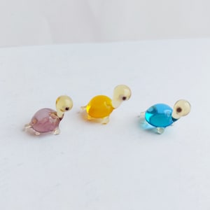 Pode incluir: Três miniaturas de tartarugas de vidro nas cores roxo, amarelo e azul. Cada tartaruga tem cabeça e pernas transparentes, com olhos escuros. As tartarugas são exibidas em uma superfície branca.