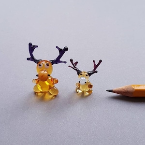 May include: Two miniature glass reindeer figurines with amber bodies and purple antlers. The reindeer are next to a yellow pencil, suggesting their small size. The figurines have detailed features and are set against a gray background.