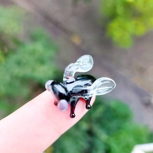 May include: A small glass sculpture of a fly with clear wings and a black body. The fly is perched on a finger.