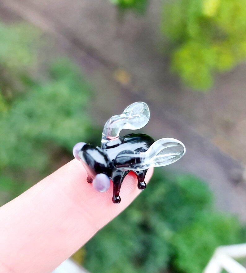May include: A small glass sculpture of a fly with clear wings and a black body. The fly is perched on a finger.