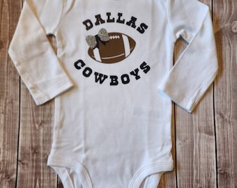 May include: White long-sleeved baby bodysuit with the text "Dallas Cowboys" and a brown football with a silver glitter bow on top.