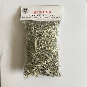 May include: A clear plastic bag filled with shredded United States currency. The bag has a white label with the text "$200.00 UNITED STATES GENUINE SHREDDED CURRENCY". The shredded currency is green and white.