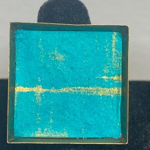 May include: Square ring with a teal and gold design. The ring has a gold-colored frame and a teal center with a horizontal gold line. The ring is displayed on a black surface.