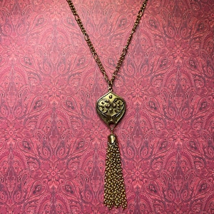 Op de afbeelding: Een goudkleurige ketting met een decoratieve hanger en kwast. De hanger heeft een sierlijk ontwerp en de kwast hangt eronder. De ketting wordt gepresenteerd tegen een roze paisley achtergrond.