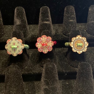 May include: Three colorful rings with floral designs. The rings are displayed on black velvet ring holders.
