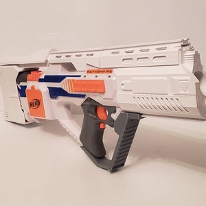 May include: White and gray Nerf Rayvenfire blaster with orange accents. The blaster has a long barrel and a grip with a textured surface. The blaster has a white and blue design on the side with the word "RAYVENFIRE" printed in black.