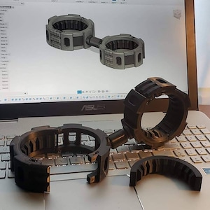 May include: Three black plastic 3D printed parts, possibly for a camera or other device, on a laptop keyboard. The parts are in various stages of assembly.